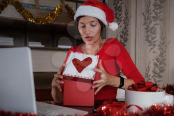 Remote Christmas video party. Family quality time during pandemic. Online video call conferencing. Young woman wearing red Santa hat and holding gift box. Virtual meeting conference calling from home