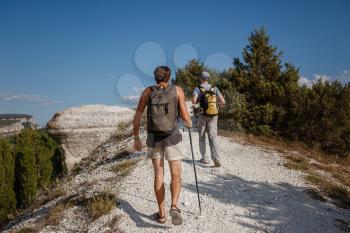 Two Men walking on rocky slope carrying Backpacks using trekking Sticks. Mountains valley View beside of People. summer travel to nature. Hiker in background.