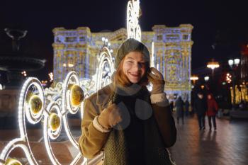 young smiling woman posing on the street. Festive Christmas fair in the background. Model wearing a stylish winter coat, knitted cap, scarf. Feeling happy in big city. Spending winter vacations Moscow