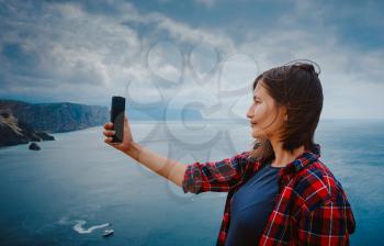 woman traveling with backpack tourist on seashore in summer. Enjoying Beautiful clouds sky among Mighty Cliffs Meeting Ocean. A woman takes a photo or video for social media