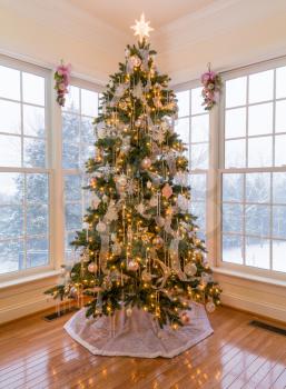 Christmas tree in modern home with snow falling outside the windows on the trees and garden