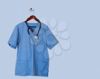 Blue medical scrubs uniform shirt hanging on a hanger with stethoscope and on hanger and hook on blue wall ready for work