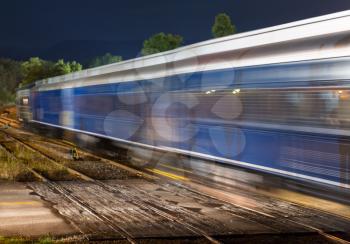 Blurred railway carriages move fast past a deserted railroad crossing with the headlights of a waiting car illuminating the train