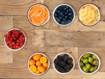 Top view of a white glass bowls of various sorts of organic fruits including, banana, orange, blueberry, redcurrant, kumquat, blackberry and kiwi berry and sitting on old wood table surface