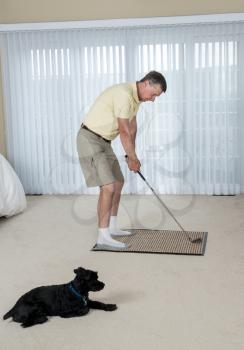 Senior caucasian man practicing his golf grip and swing in bedroom at home as pet dog looks on