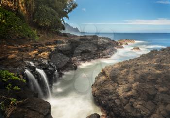 Rugged rocks around small waterfall falling into the ocean with the Na Pali coastline by Hanalei in the distance