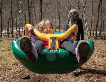 Grandma with her young granddaughter playing in a modern plastic swing in forest playground