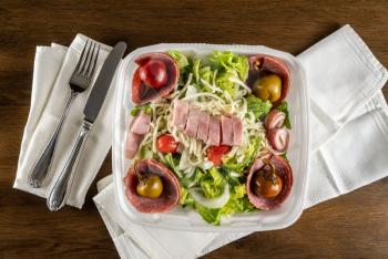 Flat lay view of antipasta salad with ham, salami peppers and lettuce in a delivery plastic box and eaten at home with napkin