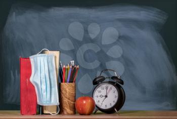 Concept for back to school with coronavirus or Covid-19 with books, apple and alarm clock against a chalkboard with face mask