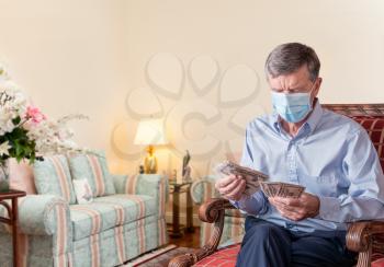 Senior caucasian man counting some US dollar bills. He is wearing face mask to prevent coronavirus. Composite into home background