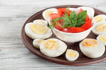 Boiled hen eggs and red tomato in a clay dish on a wooden background