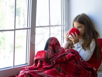The girl wrapped herself in a warm blanket sits by the window and drinks a hot drink from a cup