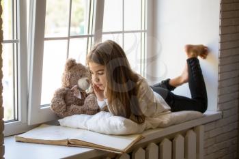 Little Girl lies on the windowsill next to her favorite toy bear cub and reads an old big book