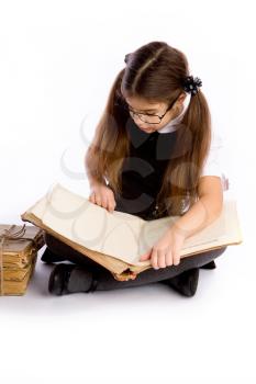 A little schoolgirl in glasses holds an enormous old book in her hands and reads it sitting on the floor