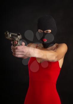 sexy girl in a tight bright red dress and balaclava holds in her hand and aims from an automatic pistol on a dark background