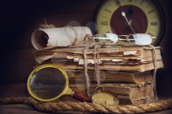 a still life with old scruffy books reading glasses, a clock and a magnifying glass on a dark background