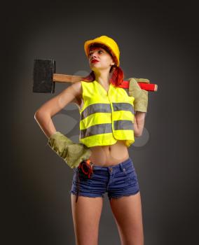 A girl in a yellow helmet mittens construction vest in shorts holding a huge hammer on a dark background