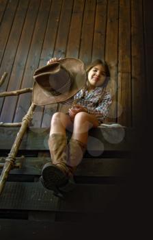 Little girl in a wide-brimmed cowboy hat and traditional dress in high boots and with a lasso posing on a dark wooden background