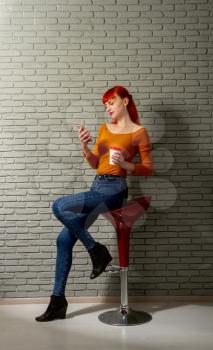 Young red-haired girl with a cup of coffee is reading something thoughtfully on her smartphone against a gray brick wall sitting on a high bar stool.