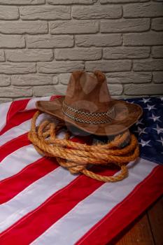 Traditional cowboy brown hat, lasso of coarse rope and a US flag against a dark brick wall