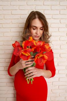 Beautiful young girl in the sixth month of pregnancy in a bright red dress with a bouquet of tulips on the background of a brick wall.