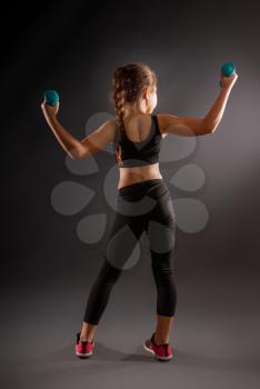 little girl in sportswear engaged with small dumbbells on a dark background