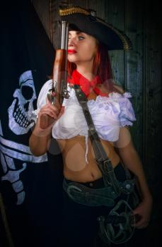 Young attractive armed girl pirate captain looks into the distance on the background of the flag Jolly Roger