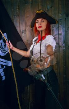 Young attractive armed girl pirate captain looks into the distance on the background of the flag Jolly Roger