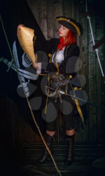 Young attractive armed girl pirate captain examines an old map on a background of the flag Jolly Roger