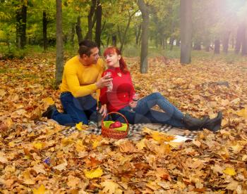 husband and wife in bright clothes for relaxing settled down on a picnic among the trees and fallen leaves with a bottle of wine and glasses.