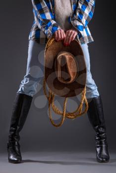 Legs in jeans and boots of a cowboy girl in a checkered shirt with a lasso and a wide-brimmed hat on a dark background