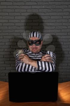 funny petty crook in exaggerated prisoner's clothes and a black mask with a silly face steals money on the Internet from gullible users through a laptop.