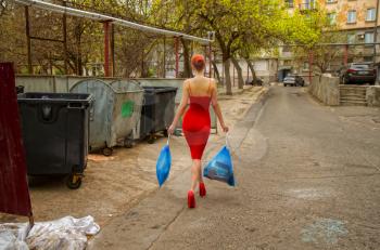 Stylish young girl in an evening dress and in red high-heeled shoes with bags of garbage found the only opportunity to walk during the virus.