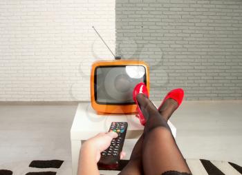 A girl sits in front of a small old TV with her legs in bright red shoes on the table and turns it on with the remote control