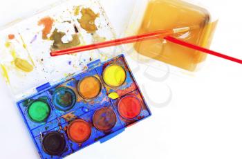 Top down view of old used watercolor palette box with paint brushes in cup with water on white paper.