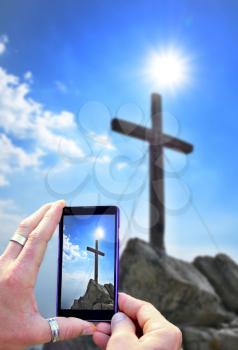Photographing and taking photo of wooden cross on peak by smart phone. View over smart phone screen during photographing of landscape shot.