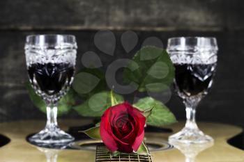 Red Rose and Wine Glasses Resting On Acoustic Guitar With Wooden Background