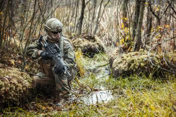 Norwegian Rapid reaction special forces FSK soldier sitting on the swamp. Field camo uniforms, combat helmet and eye-wear goggles are on