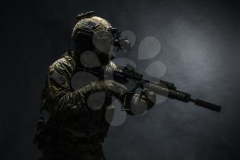 Bearded soldier in Combat Uniforms with weapon, plate carrier and combat helmet are on. Studio shot, dark background