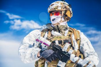 Army serviceman in winter camo somewhere in the Arctic. He wears chest rig and huge backpack, suffers from the extreme cold and strong wind, but endures while mission continues. Blue sky background