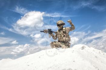 Airsoft military games player in camouflage uniform and helmet armed assault rifle replica, sitting on knee on top of sand dune with sky on background and showing freeze hand signal to his teammates