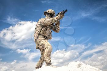 War game player in military camouflage uniform, equipped with tactical ammunition, aiming with service rifle replica while moving in sand with cloudy sky on background, during airsoft game event