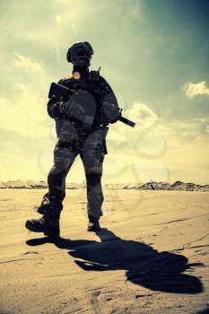 Special operations forces soldier in camouflage combat uniform, helmet and glasses walking in desert with service rifle in hands low angle view. War conflict, military campaign in Middle East region