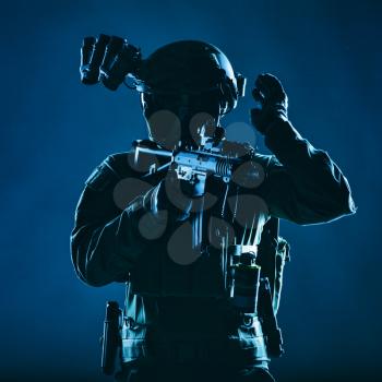 Special operations soldier, SWAT team fighter in mask and glasses, equipped night vision device, armed short barrel service rifle, looking back and showing freeze hand signal, low key studio shoot