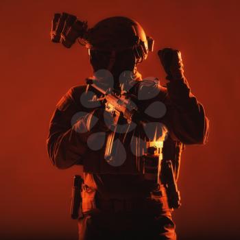 Special operations soldier, SWAT team fighter in mask and glasses, equipped night vision device, armed short barrel service rifle, looking back and showing freeze hand signal, low key studio shoot