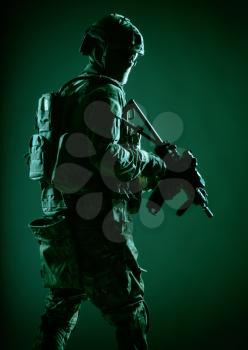 Army soldier in helmet, battle uniform with tactical ammunition, hiding face behind mask and glasses, looking back over shoulder, sneaking in darkness with service rifle in hand, low key, studio shoot