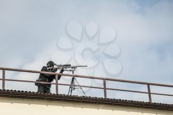 Police special operations or security team sniper standing on roof of city building and aiming with telescopic optical sight on sniper rifle mounted on tripod. SWAT shooter engage long range targets