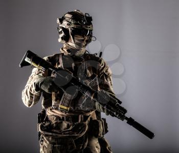 Special forces fighter in battle uniform and helmet with radio headset, face mask and ballistic glasses, standing with equipped laser sight and silencer service rifle studio portrait isolated on white