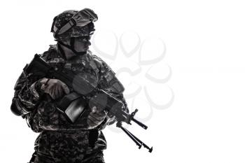 Low key side portrait of US Army Special Forces soldier in military camouflage uniform protected with helmet, body armour, holding machine gun desaturated, isolated on white background with copyspace