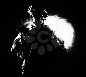 Low key studio portrait of private security service contractor, army infantry rifleman, US marine raider in helmet, sunglasses, camouflage uniform posing with weapon on black background with backlight